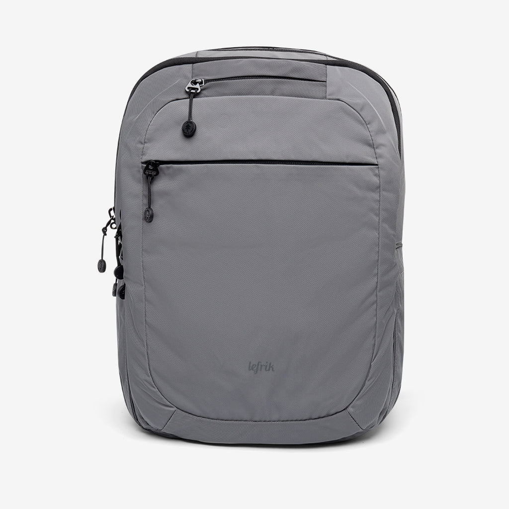 Mochila gris claro de diseño minimalista, con un bolsillo frontal con cremallera, una cremallera lateral y el nombre de la marca "Lefrik" sutilmente impreso cerca de la parte inferior. El fondo es blanco liso.