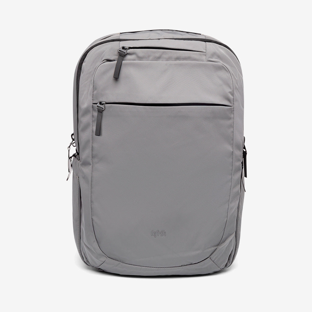 Una mochila gris claro de diseño minimalista, con dos bolsillos delanteros con cremallera y cremalleras laterales, sobre un fondo blanco liso.