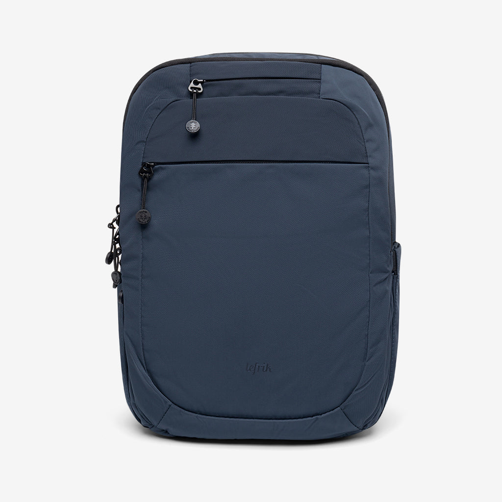 Una mochila azul marino de diseño minimalista, con un bolsillo frontal con cremallera, cierre superior de cremallera y un pequeño logotipo en la parte inferior central delantera, sobre un fondo blanco liso.